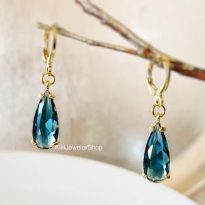 May include: Two gold-plated leverback earrings, each with a teal, teardrop-shaped gemstone. The earrings hang from a branch, highlighting their design. The earrings are a jewellery item.