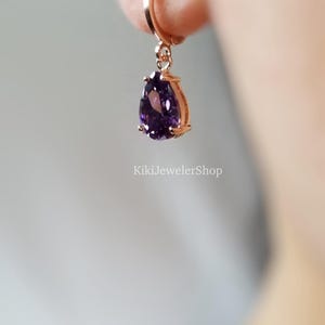 Op de afbeelding: Rosegouden oorbel met een dieppaarse edelsteen in druppelvorm. De oorbel hangt aan een kleine oorring. De tekst "KikiJewelerShop" is zichtbaar op de achtergrond.