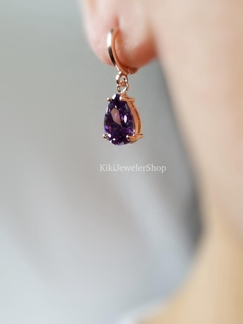 Op de afbeelding: Rosegouden oorbel met een dieppaarse edelsteen in druppelvorm. De oorbel hangt aan een kleine oorring. De tekst "KikiJewelerShop" is zichtbaar op de achtergrond.