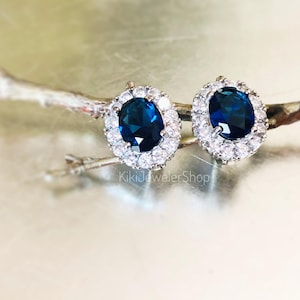 May include: A pair of oval-shaped stud earrings with a dark blue center stone surrounded by a halo of smaller, clear stones. The earrings are set on a silver-colored metal and are displayed on a thin, brown twig against a light background. The text "KikiJewelerShop" is visible.