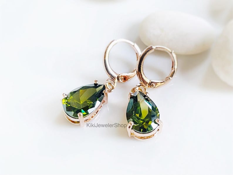 Op de afbeelding: Rosegouden oorringen met peer-vormige olijfgroene edelstenen. De oorbellen worden gepresenteerd op een witte ondergrond. De oorbellen zijn van KikiJewelerShop.