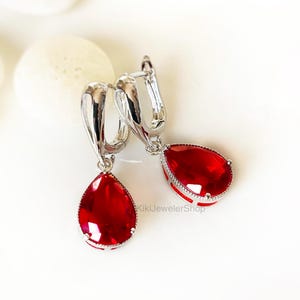 Puede incluir: Un par de pendientes colgantes de tono plateado con gemas rojas en forma de lágrima. Cada pendiente tiene una parte superior de aro de metal pulido con cierre de bisagra y un engaste de piedra roja facetada. El texto KikiJewelerShop es visible.