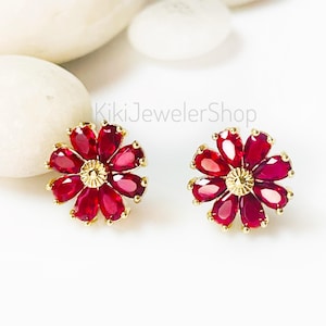 May include: A pair of flower-shaped stud earrings. Each earring features a gold-colored base with a central gold accent and petals of deep red gemstones. The earrings are set against a white background with smooth, white stones.