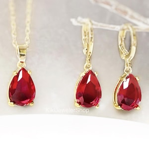 May include: A gold jewelry set consisting of a necklace with a pear-shaped red gemstone pendant and a pair of matching drop earrings. Each piece features a faceted deep red stone set in a gold-toned metal frame. The text KikiJewelerShop is visible.