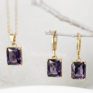 May include: A gold-toned jewelry set featuring a necklace and a pair of earrings, each with a rectangular, deep purple gemstone. The necklace pendant and earring drops are framed in gold. The shop name "KikiJewelShop" is visible.