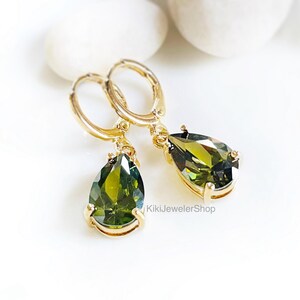 Op de afbeelding: Goudkleurige oorringen met een peer-vormige, olijfgroene edelsteen. De oorbellen worden gepresenteerd op een wit oppervlak, met de winkelnaam "KikiJewelerShop". De edelstenen hebben een facetgeslepen.
