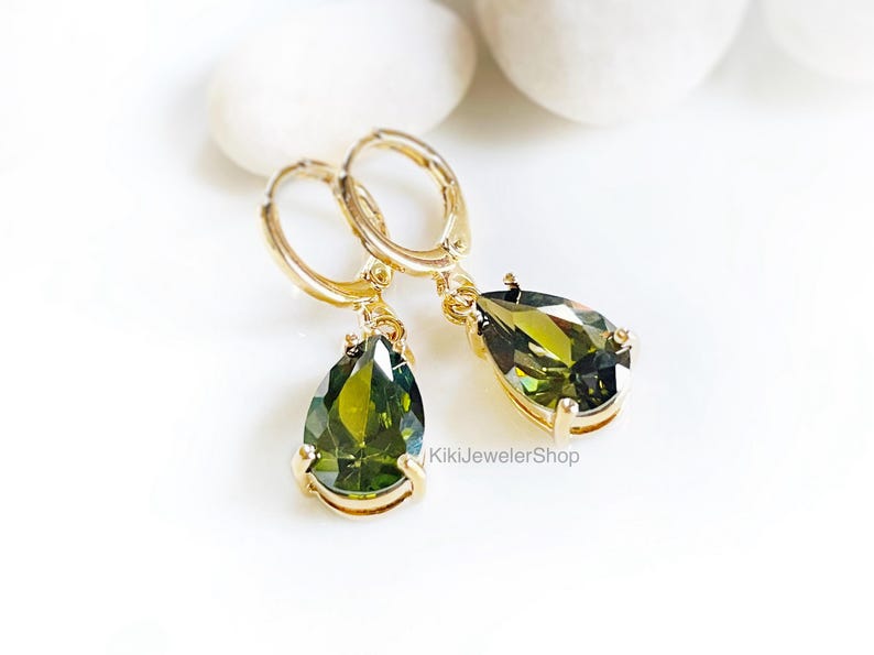 Op de afbeelding: Goudkleurige oorringen met een peer-vormige, olijfgroene edelsteen. De oorbellen worden gepresenteerd op een wit oppervlak, met de winkelnaam "KikiJewelerShop". De edelstenen hebben een facetgeslepen.