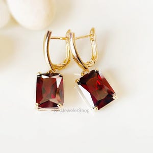May include: A pair of gold-colored earrings with rectangular, deep red gemstones. The earrings have a lever-back closure and are displayed on a white surface. The gemstones are a rich, dark red color.