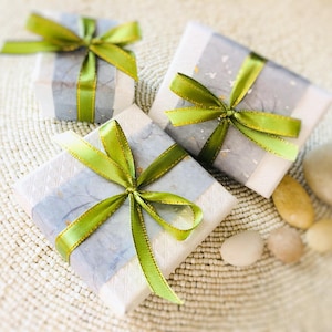 Op de afbeelding: Drie ingepakte geschenkdozen met groene en gouden strikken. De dozen zijn verpakt in wit en grijs papier met een subtiel patroon. Verschillende gladde, lichtgekleurde stenen zijn in de buurt van de cadeaus op een geweven oppervlak geplaatst.