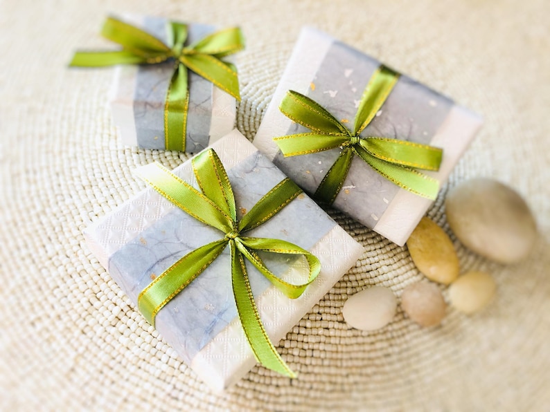 Op de afbeelding: Drie ingepakte geschenkdozen met groene en gouden strikken. De dozen zijn verpakt in wit en grijs papier met een subtiel patroon. Verschillende gladde, lichtgekleurde stenen zijn in de buurt van de cadeaus op een geweven oppervlak geplaatst.