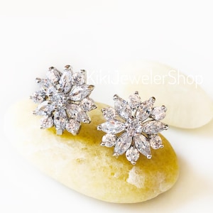 May include: A pair of silver-toned stud earrings with a floral design. Each earring features a central, round, clear stone surrounded by marquise-cut clear stones, creating a flower-like shape. The earrings are displayed on a light yellow stone.
