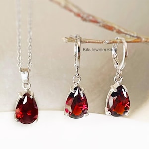 May include: A jewelry set featuring a silver necklace with a teardrop-shaped, deep red gemstone pendant, and matching earrings. The earrings are hoop style with the same gemstone. The gemstones have a faceted cut, reflecting light.
