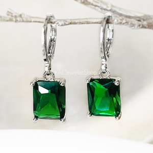 May include: A pair of silver-toned leverback earrings with emerald-cut, vibrant green gemstones. The earrings are displayed on a light-colored, neutral background. The text "KikiJewelerShop" is visible.