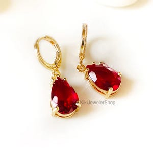 May include: A pair of gold-colored hoop earrings with teardrop-shaped, deep red gemstones. The earrings are displayed on a white surface. The text "KikiJewelerShop" is visible on the image.