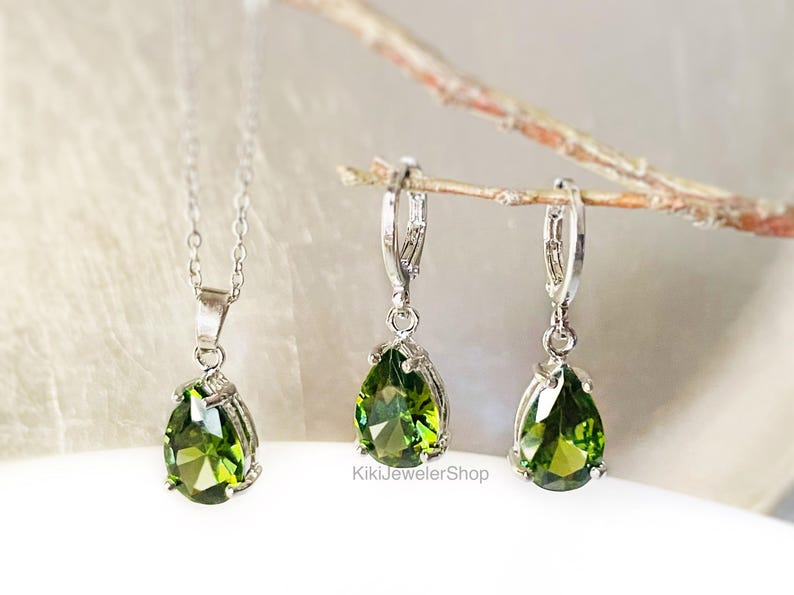 Op de afbeelding: Een zilveren ketting met een peervormige groene edelsteen hanger en een paar bijpassende oorbellen. De oorbellen en hanger zijn gezet in zilver metaal. De sieraden worden gepresenteerd op een lichte achtergrond.