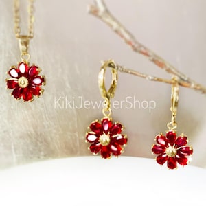 May include: A gold-toned jewelry set featuring a necklace and a pair of earrings, each adorned with a red and white flower design. The floral pendants and earrings are set against a soft, blurred background. The text "KikiJewelerShop" is visible.