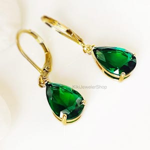 May include: A pair of gold leverback earrings featuring faceted, pear-shaped emerald green stones. The earrings are set in a gold-toned metal frame with a secure clasp mechanism. The text KikiJewelerShop is visible in the center of the image.
