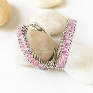 May include: A pair of silver-toned hoop earrings featuring a row of square-cut pink gemstones set along the front. The earrings have a hinged closure mechanism and the text KikiJewelerShop is visible on the inner surface of one hoop.