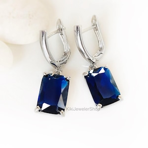 May include: A pair of silver-toned drop earrings featuring rectangular, deep blue gemstones. The earrings have a lever-back closure and are set against a white background. The gemstones are a striking, saturated color.