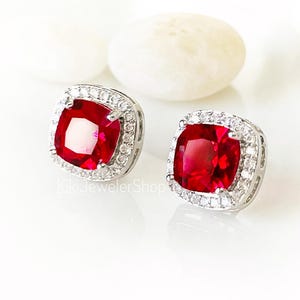 May include: A pair of stud earrings featuring a square-cut, vibrant red gemstone at the center, surrounded by a halo of small, clear white stones set in a polished silver-toned metal frame. The text KikiJewelerShop is visible at the bottom.