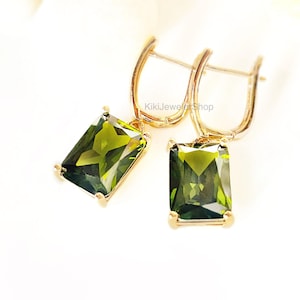 May include: A pair of gold-colored drop earrings with rectangular, olive-green gemstones. The earrings have a lever-back closure and are set against a white background. The text "KikiJewelerShop" is visible.