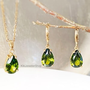 Op de afbeelding: Een goudkleurige sieradenset met een ketting en oorbellen. Elk stuk toont een peervormige, levendige groene edelsteen. De hanger van de ketting en de oorbelhangers zijn opgehangen aan delicate gouden kettingen en haken. De sieraden worden gepresenteerd op een wit oppervlak.