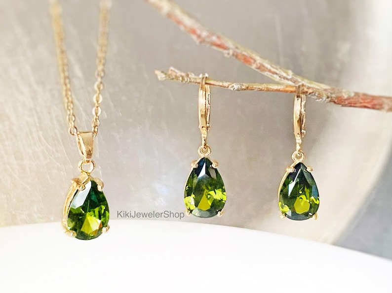 Op de afbeelding: Een goudkleurige sieradenset met een ketting en oorbellen. Elk stuk toont een peervormige, levendige groene edelsteen. De hanger van de ketting en de oorbelhangers zijn opgehangen aan delicate gouden kettingen en haken. De sieraden worden gepresenteerd op een wit oppervlak.