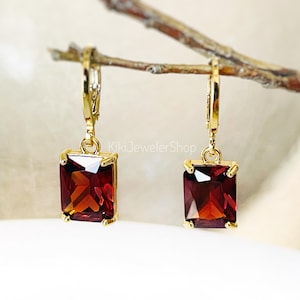 May include: A pair of gold-colored leverback earrings with rectangular, deep red gemstones. The earrings are suspended from a branch, showcasing their design and color. The earrings are from KikiJewelerShop.