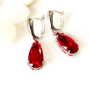 May include: A pair of silver-toned drop earrings with large, teardrop-shaped, ruby-red gemstones. The earrings have a lever-back closure and are set against a white background. The gemstones are faceted, catching the light.