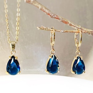 Puede incluir: Un conjunto de joyas doradas que incluye un collar y un par de pendientes. Cada pieza presenta una piedra preciosa azul oscuro en forma de lágrima. El colgante del collar y los colgantes de los pendientes cuelgan de cadenas y aros dorados. Las joyas se exhiben sobre un fondo claro.