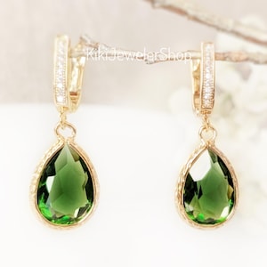 May include: A pair of gold drop earrings featuring faceted pear-shaped green stones suspended from hinged hoops. The hoops are accented with small clear stones, and the teardrop-shaped green gems are set in gold frames with a textured edge detail.