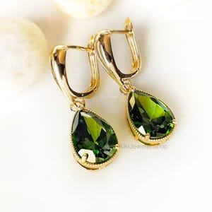 May include: Gold-tone leverback earrings with pear-shaped, faceted, green gemstones. The gemstones are set in a gold-tone bezel with a delicate beaded edge. The earrings are displayed on a white surface with a few white stones.