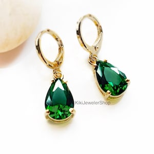 Può includere: Un paio di orecchini dorati con chiusura a leva e pietre preziose verde smeraldo a forma di goccia. Gli orecchini sono presentati su una superficie bianca, con il testo "KikiJewelerShop" visibile.