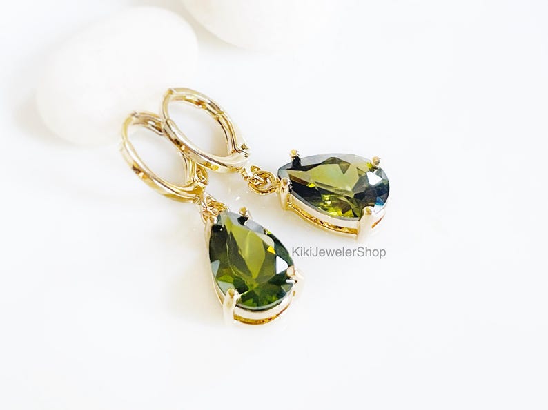 Op de afbeelding: Goudkleurige oorhangers met olijfgroene, peervormige edelstenen. De oorbellen hebben een klapsluiting en worden gepresenteerd op een wit oppervlak. De edelstenen hebben meerdere facetten. KikiJewelerShop.