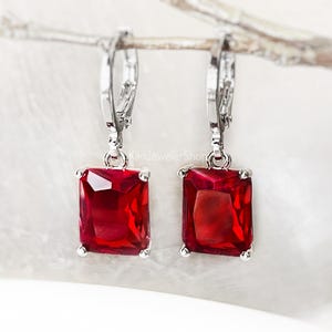May include: A pair of silver-toned leverback earrings with large, rectangular, deep red gemstones. The gemstones are set in a four-prong setting, and the earrings are displayed against a soft, neutral background. The earrings are a classic jewelry design.