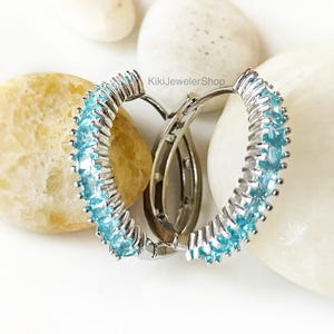 May include: Silver hoop earrings with a row of light blue gemstones. The earrings are displayed on a white surface with smooth, light-colored stones. The earrings are a jewelry item.