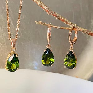 Op de afbeelding: Een set van een ketting en oorbellen in ros&eacute;goud met peervormige groene edelstenen. De ketting heeft een fijne ketting en de oorbellen zijn van het creoolse type. De sieraden worden op een tak gepresenteerd.