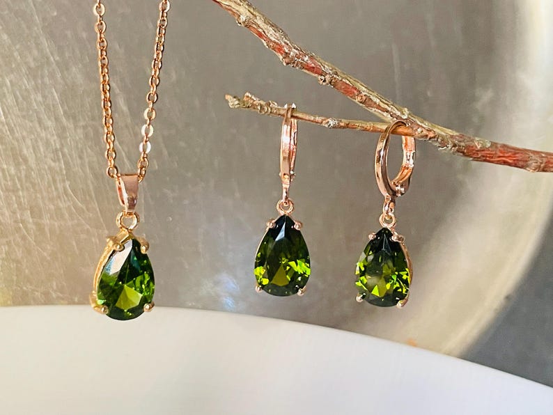 Op de afbeelding: Een set van een ketting en oorbellen in ros&eacute;goud met peervormige groene edelstenen. De ketting heeft een fijne ketting en de oorbellen zijn van het creoolse type. De sieraden worden op een tak gepresenteerd.