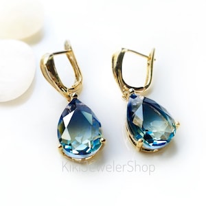 May include: Gold-coloured drop earrings featuring pear-shaped, faceted blue-green gemstones. The earrings have a lever-back closure and are displayed on a white surface. The text "KikiJewelerShop" is visible.