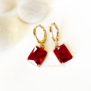 Könnte beinhalten: Goldfarbene Creolen mit rechteckigen, tiefroten Edelsteinen. Die Ohrringe haben einen Klappverschluss und sind vor einem weißen Hintergrund mit dekorativen weißen Steinen platziert. KikiJewelerShop.