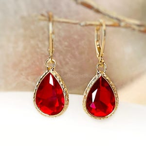 Op de afbeelding: Een paar goudkleurige oorhangers met druppelvormige, gefacetteerde rode edelstenen. De edelstenen zijn gezet in een goudkleurig montuur met een getextureerde rand. De oorbellen hebben een klapsluiting.