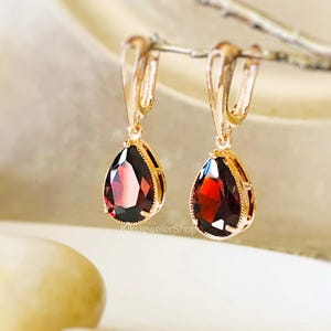 May include: A pair of gold-coloured earrings featuring teardrop-shaped, deep red gemstones. The earrings have a lever-back closure and are set in a decorative gold frame. The gemstones are faceted, catching the light.