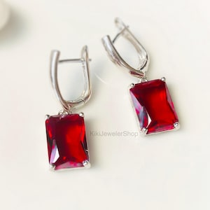 May include: A pair of silver-toned leverback earrings with rectangular, deep red gemstones. The gemstones are set in a silver frame and have a faceted appearance. The earrings are displayed on a white surface.