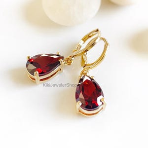 May include: Gold-colored hoop earrings with teardrop-shaped, deep red gemstones. The earrings have a lever back closure and are set in a gold-colored metal. The gemstones are faceted, catching the light. The earrings are displayed on a white surface.