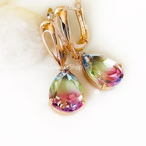 Pode incluir: Um par de brincos pendurados dourados com grandes pedras preciosas em forma de gota. As pedras preciosas exibem um gradiente de cores, incluindo azul, verde, rosa e roxo. Os brincos estão sobre um fundo branco.