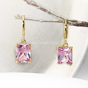 May include: A pair of gold-colored earrings with rectangular, pink gemstones. The earrings are hanging from a thin, light-colored branch. The gemstones have a faceted appearance, reflecting light. The earrings are a jewelry item.