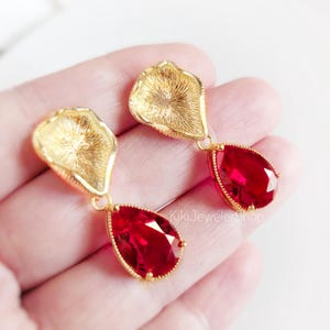May include: A pair of gold-toned drop earrings featuring a textured, petal-shaped top component connected to a faceted, pear-shaped red crystal pendant. The earrings are finished with a gold-colored metal setting and include the text KikiJewelerShop.