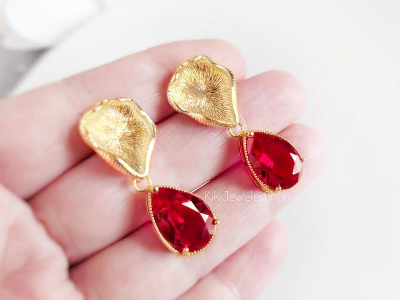 May include: A pair of gold-toned drop earrings featuring a textured, petal-shaped top component connected to a faceted, pear-shaped red crystal pendant. The earrings are finished with a gold-colored metal setting and include the text KikiJewelerShop.