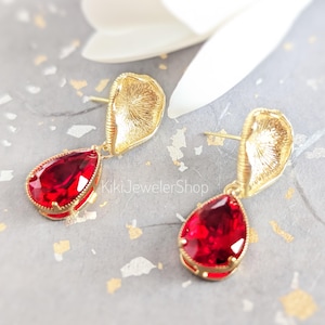 May include: A pair of gold-toned earrings featuring a textured, shell-like top component and a faceted, pear-shaped red crystal drop. The text KikiJewelerShop is visible between the earrings. The design includes a post-back closure mechanism.