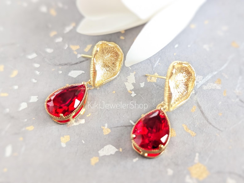May include: A pair of gold-toned earrings featuring a textured, shell-like top component and a faceted, pear-shaped red crystal drop. The text KikiJewelerShop is visible between the earrings. The design includes a post-back closure mechanism.